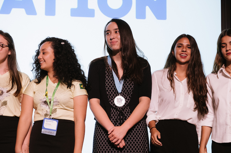Technovation Girls 2019 Team Uproot wins second place at Technovation World Pitch – a line of young women stand on a stage, the young woman in the center, Team Uproot, wears a medal around her neck